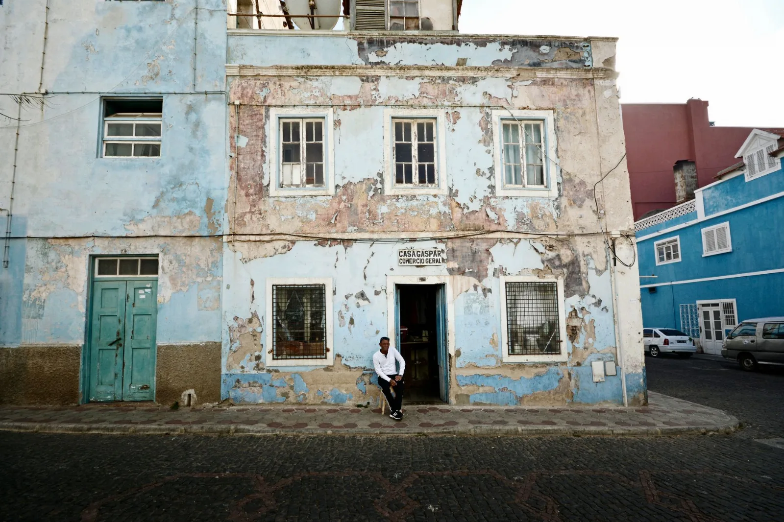 Façade bleue écaillée d'une rue en pente de Mindelo, avec un homme en chemise blanche assis sur le seuil