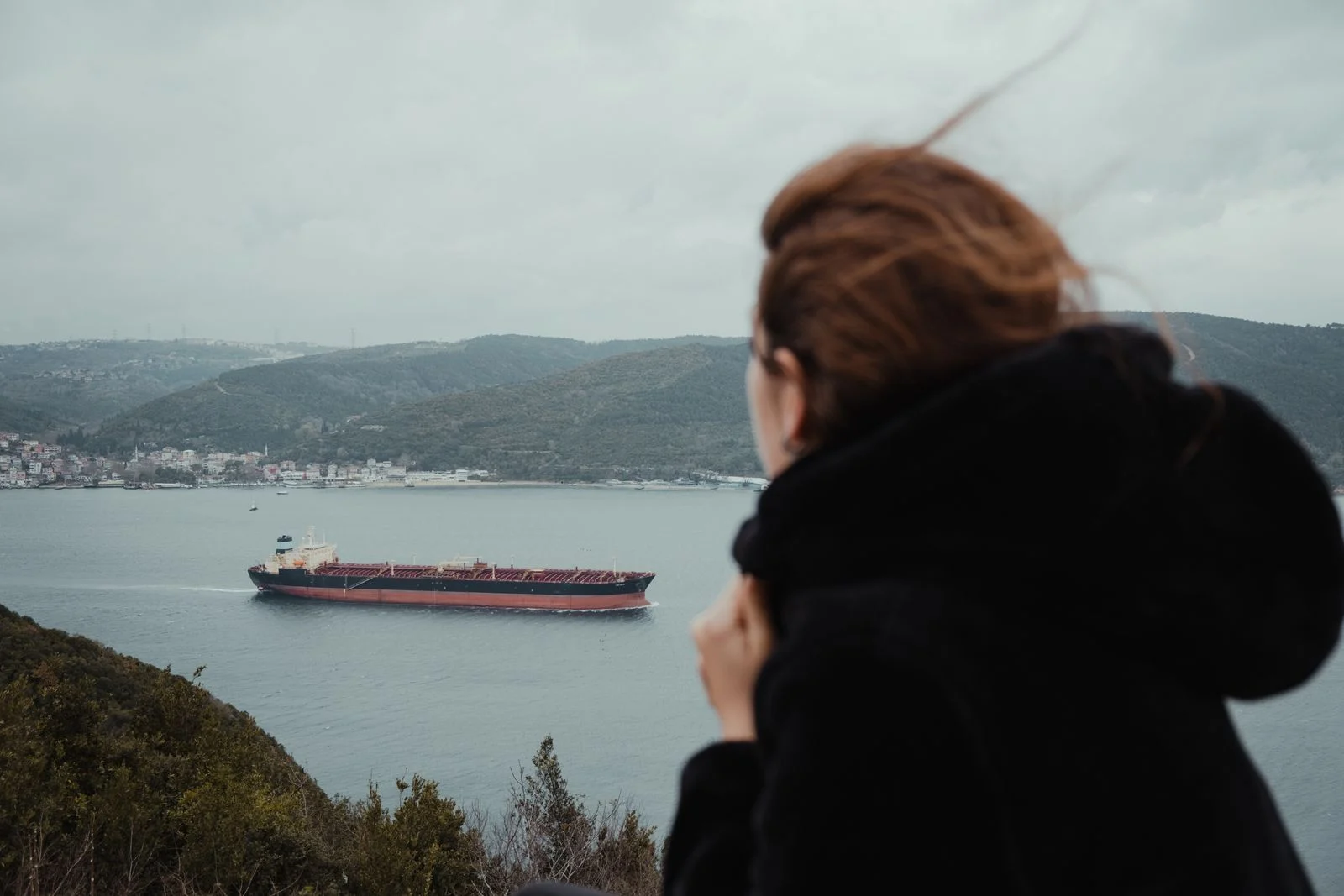 Voyageuse en manteau sombre regardant un navire depuis la côte par temps couvert