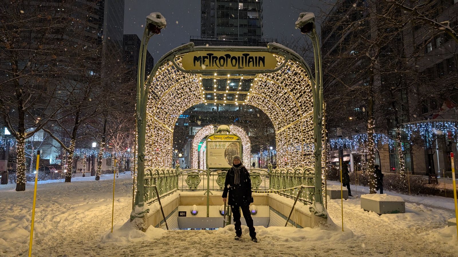 Entrée de métro de style Métropolitain illuminée la nuit sous la neige, à Montréal, un passant emmitouflé devant