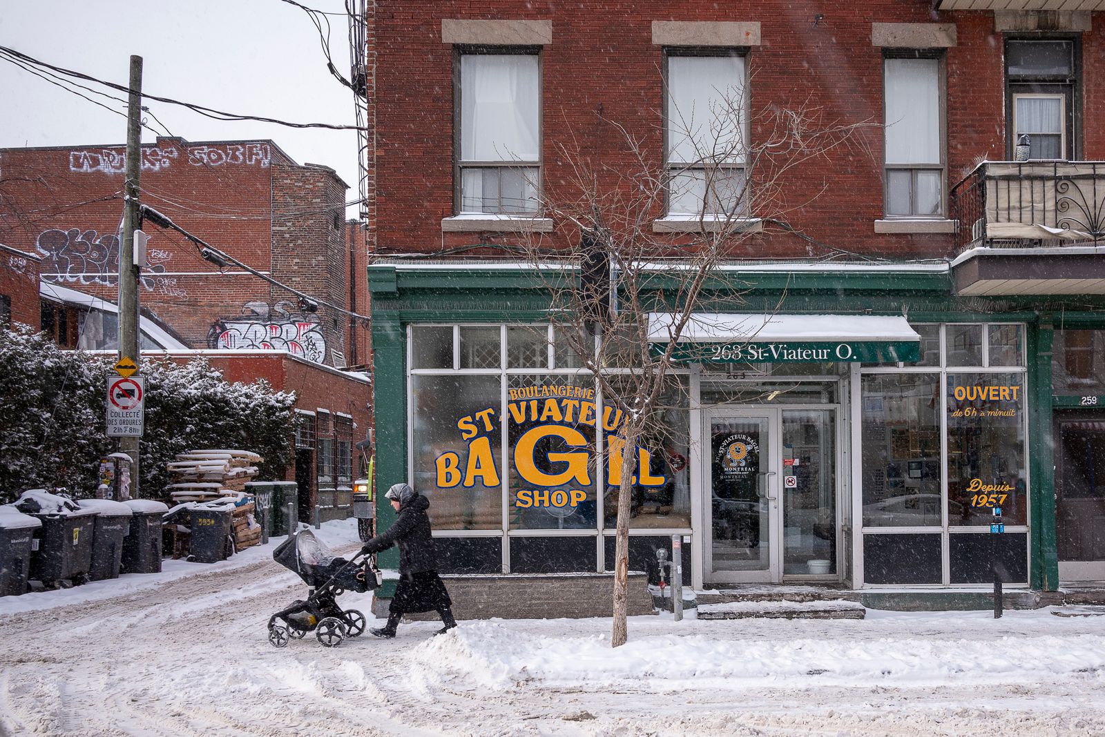 Rue enneigée de Montréal devant une boulangerie de bagels, une passante en manteau d'hiver pousse une poussette