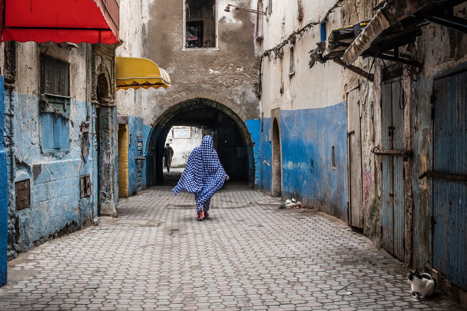 Une femme de dos, drapée d'un voile bleu à pois, dans une ruelle pavée d'Essaouira