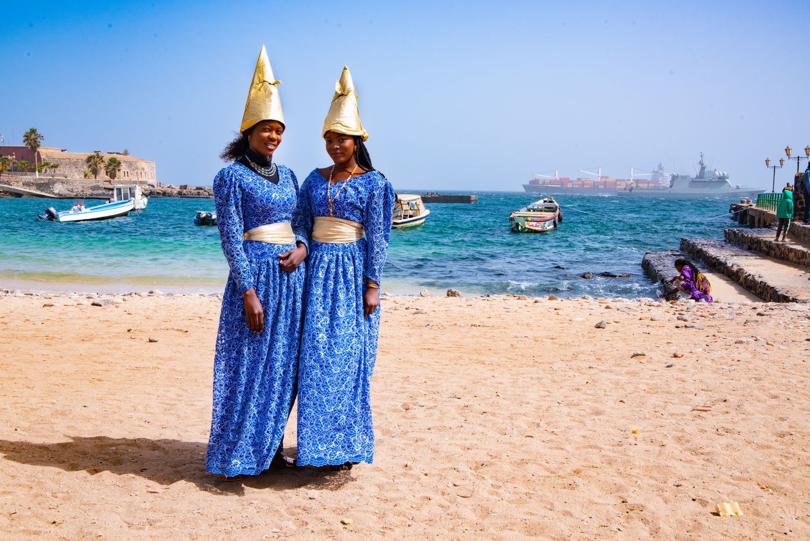 Deux femmes en robes longues bleues à motifs et coiffes coniques dorées, debout sur une plage de Dakar