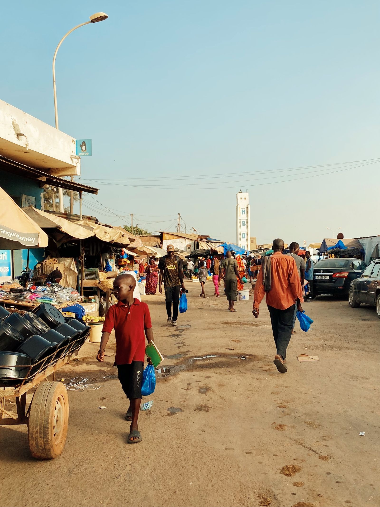 Rue de sable bordée d'étals à M'bour, avec des passants en t-shirt et pantalon