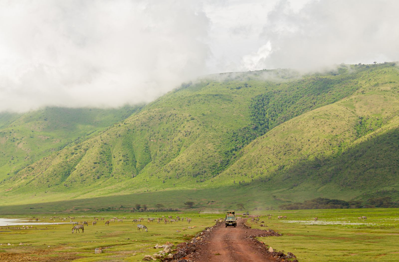 Véhicule de safari sur une piste rouge au fond du cratère du Ngorongoro, sous des parois vertes coiffées de nuages